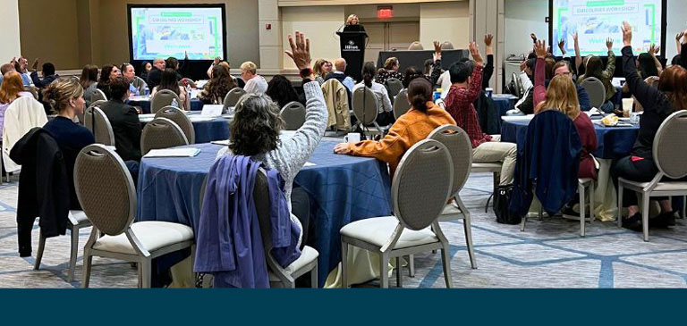 Leadership_Workshops_768x365 Conference room with people at various tables and some are holding up their hands.