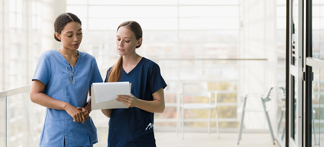 Two nurses consulting over paperwork in a hallway.